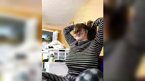 Media: Video of a woman with light skin, brown hair, wearing a black and white striped sweater, sitting on a couch with crossed arms, in a laundry room with a white washer and dryer, and a TV in the background.