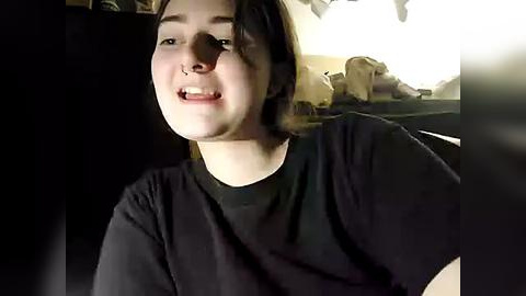 Media: Video of a young woman with light skin, brown hair, and a black t-shirt, smiling, in a dimly lit room with a messy background.