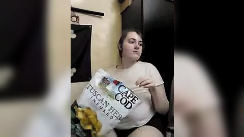 Media: A video of a plus-sized woman with dark hair, wearing a white t-shirt, sitting on a toilet, holding a bag of Cabbage Patch Kids and eating Doritos.