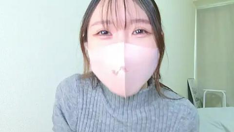 Media: A video of an Asian woman with light skin and straight black hair, wearing a gray ribbed sweater and a white surgical mask, standing indoors with a plain white wall background.