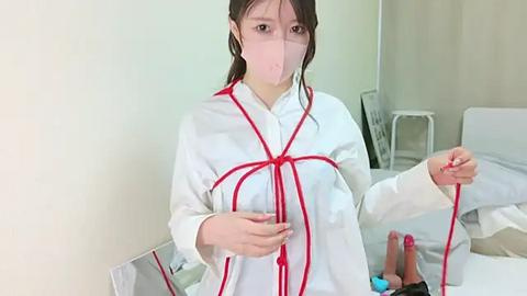 Media: A video of a young Asian woman in a white lab coat with red ropes, holding a phallus-shaped object, standing in a sterile hospital room with a white bed and medical equipment.