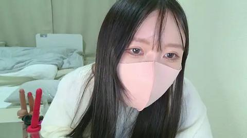 Media: Video of an Asian woman with long black hair and fair skin, wearing a pink surgical mask, standing in a softly lit room with beige walls and a bed with white sheets in the background.