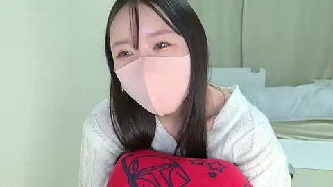 Media: Video of an Asian woman with long black hair, wearing a pink face mask and a white knitted sweater, holding a red pillow with black star patterns. Background shows a white bed and beige wall.