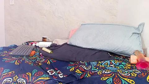 Media: Video of a messy bed with a blue and multicolored patterned comforter, a laptop, a keyboard, a white mug, and a dark shirt on a white textured wall.