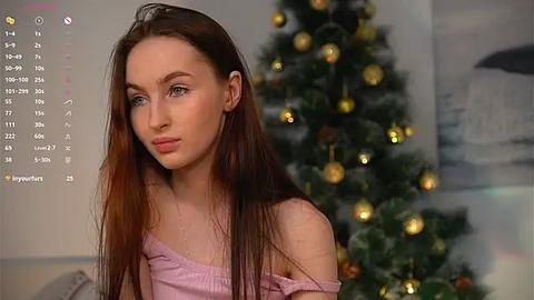 Media: Video of a young woman with long brown hair, light skin, and blue eyes, wearing a pink off-shoulder top, standing in front of a decorated Christmas tree with gold ornaments and fairy lights, in a cozy indoor setting.