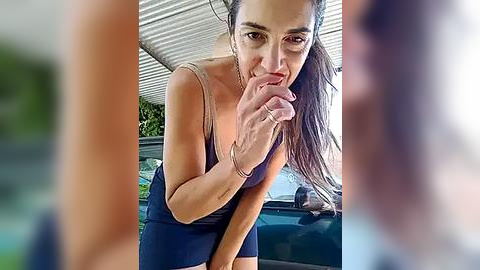 Media: Video of a dark-haired woman with a slender build, wearing a sleeveless top and short skirt, playfully biting her finger under a carport.