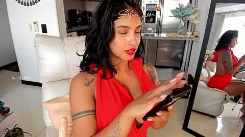 Media: Video of a tattooed woman with medium skin tone and long black hair, wearing a red sleeveless top, applying makeup in a modern living room with white leather couches and a mirror reflecting her back.