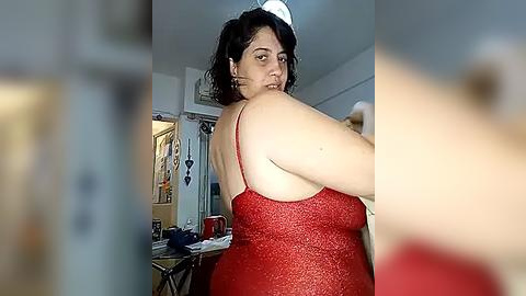 Media: A video of a plus-sized woman in a red glitter dress and white cap, standing in a cluttered bedroom with a bed and shelves visible.