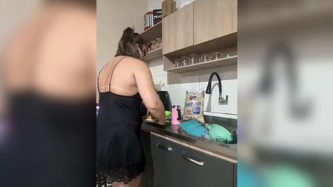 Media: Video of a woman with light brown skin and shoulder-length hair in a black lace lingerie dress washing dishes in a modern kitchen with wooden cabinets, a stainless steel sink, and white tiled backsplash.