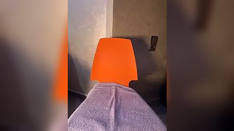 Media: A video of a modern chair with a bright orange backrest and a purple cloth draped over it, situated against a muted beige wall with a textured surface. The chair is centrally positioned in a dimly lit room.