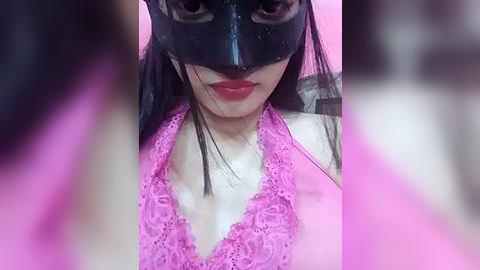 Media: Video of a woman with long black hair, wearing a black mask and a pink lace bralette, set against a pink background, with blurred figures in the foreground.