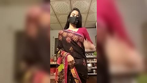 Media: Video of a woman with dark skin, black mask, and black sari with colorful floral patterns, standing in a kitchen with a white ceiling and tiled backsplash.