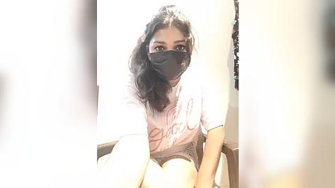 Media: Video of a young woman with long black hair, wearing a white T-shirt, grey shorts, and a black mask, sitting indoors with sunlight creating a halo effect.