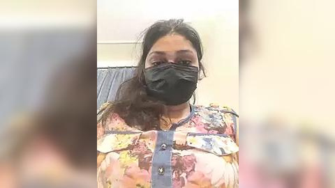 Media: Video of a young woman with medium brown skin, wearing a floral blouse, black face mask, and denim overalls, seated indoors with a blurred background.