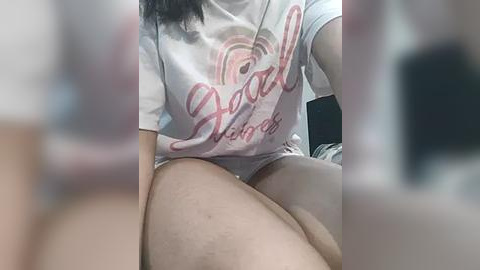 Media: A video of a person with light skin, wearing a white \"Hello Kitty\" t-shirt, sitting on a bed. The image is blurred, with a smartphone's camera lens visible in the foreground.