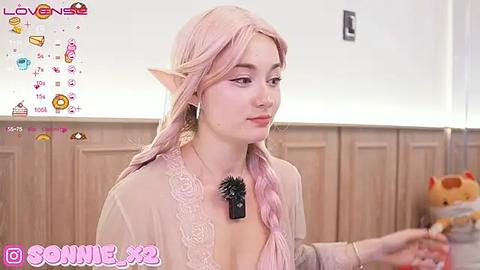 Media: Video of a fair-skinned, pink-haired, elf-like woman with pointy ears, wearing a sheer, lace-trimmed top, holding a stuffed bear. Background shows a wooden-paneled wall and a \"Love Games\" social media overlay.