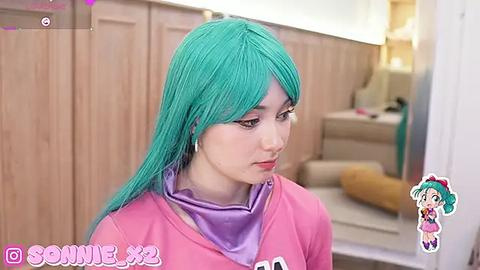 Media: Video of a young woman with teal hair, wearing a pink hoodie and purple scarf, standing indoors against wooden cabinets.