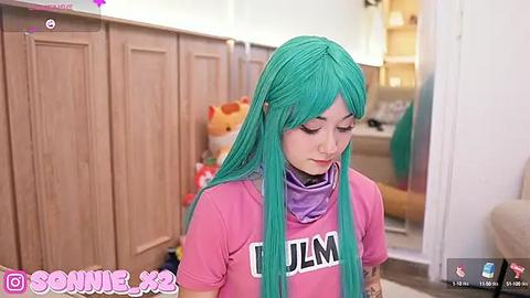 Media: Video of a young woman with long, vibrant turquoise hair, wearing a pink t-shirt and a purple mask, standing in a cozy room with wooden paneling, plush toys, and a plushie tiger.