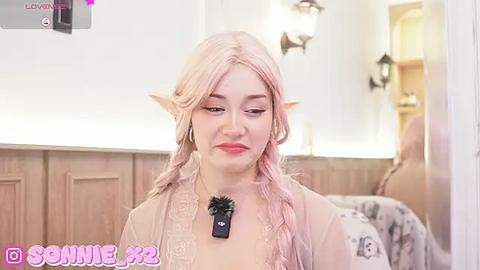 Media: Video of a fair-skinned woman with long, pale pink hair and pointed elf ears, wearing a sheer, light pink top, standing in a cozy, warmly lit room.