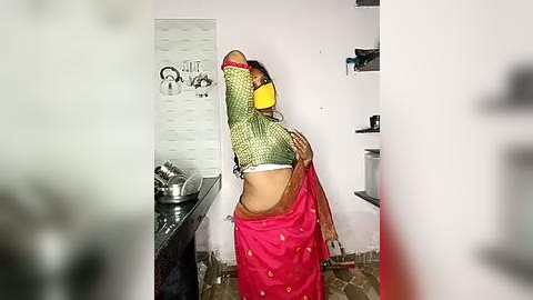 Media: Video of a woman in a yellow headscarf, pink saree, and black face mask, stretching in a small, cluttered kitchen with white tiled walls, a sink, and a stove.