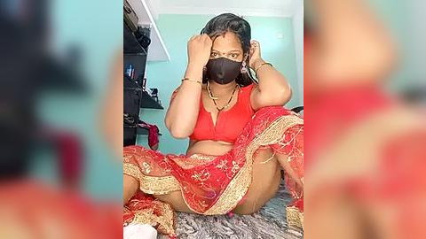 Media: Video of a South Asian woman in a red saree, sitting on a grey rug, fixing her hair, wearing a face mask, in a modestly furnished room.