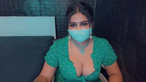 Media: A video of a South Asian woman with medium skin tone and dark hair, wearing a green blouse with a deep neckline, revealing cleavage, a blue mask, and a silver necklace, seated on a bed with dark curtains in the background.