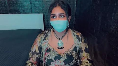 Media: Video of a South Asian woman with dark skin and curly hair, wearing a floral blouse, turquoise face mask, and silver jewelry. She sits on a bed with a dark headboard against a dark, textured wall.