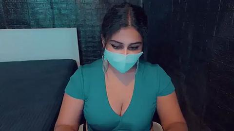 Media: Video of a Latina woman with medium skin tone, dark hair, wearing a teal V-neck top, a green face mask, and dangling earrings, seated on a black couch against a dark brick wall.