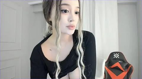 Media: Video of an Asian woman with long, wavy, blonde hair, wearing black clothing, standing indoors. She has a small tattoo on her left shoulder and a gaming chair with orange accents is visible in the background.