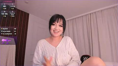 Media: Video of a smiling East Asian woman with short black hair, wearing a light gray cardigan, sitting on a bed in a modern, dimly lit bedroom with white curtains.