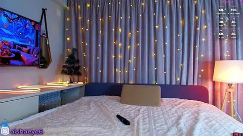 Media: Video of a modern, cozy bedroom with a TV mounted on the wall, white bedding, a beige pillow, and a floor lamp. Soft yellow fairy lights adorn the curtains, creating a warm ambiance.