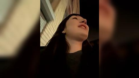 Media: Video of a young woman with long black hair, smiling upward, standing in front of a beige brick building at night.