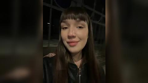 Media: Video of a young woman with long, straight, dark brown hair and blunt bangs, smiling, wearing a black leather jacket. The background is dimly lit, suggesting an indoor setting, possibly a nightclub.