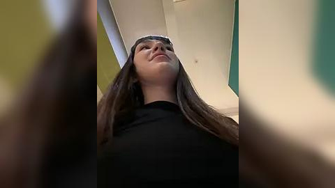 Media: Video of a young woman with long, dark hair, wearing a black shirt, standing indoors, looking up. The background is a mix of green and yellow walls.