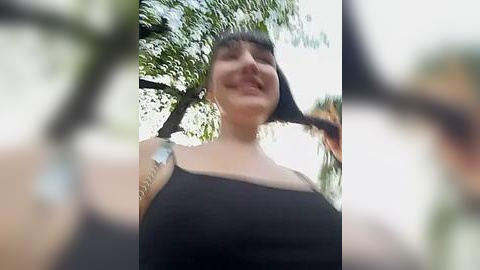 Media: A video of a smiling, light-skinned woman with dark hair, wearing a black tank top, taken from a low angle, with a blurred background of green trees.