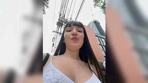 Media: Video of an Asian woman with long black hair, wearing a white lace bralette, pouting playfully, standing in a city street with power lines and buildings in the background.