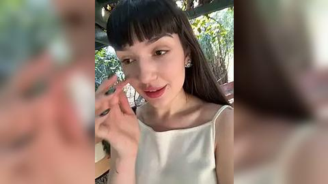 Media: Video of a young woman with straight, dark hair and bangs, wearing a white sleeveless dress, applying makeup with a brush. Background features blurred greenery and a wooden structure.