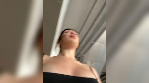 Media: Video of a woman with light skin and dark hair, wearing a black strapless dress, captured from a low angle, with blurred background showing white walls and a ceiling.