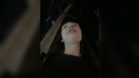 Media: A dimly lit video captures a young woman with short black hair and fair skin, lying on her back in a dark room, looking upwards. She wears a black top.