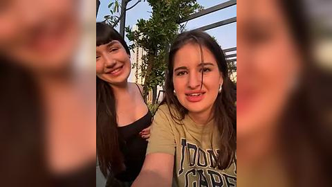 Media: Video of two young women smiling, one with long dark hair, wearing a black top, and the other with long brown hair, wearing a green t-shirt. They are outdoors, with blurred background foliage and a clear sky.