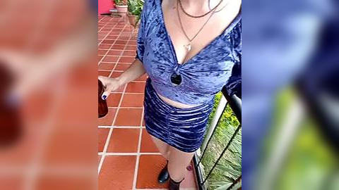 Media: Video of a fair-skinned woman with a tattoo on her thigh, wearing a blue velvet halter top and short skirt, holding a drink, on a red-tiled balcony.