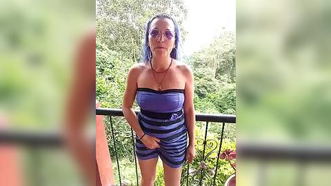 Media: Video of a woman with medium skin tone, wearing sunglasses and a strapless, blue striped romper, standing on a balcony overlooking lush greenery.