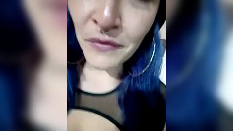 Media: Video of a young woman with pale skin, blue hair, and a septum piercing, wearing a black mesh top. Blurred background.