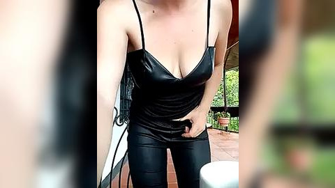 Media: Video of a fair-skinned woman with a slender build, wearing a black, low-cut, shiny leather camisole and tight black pants, standing on a terrace with a blurred green background.