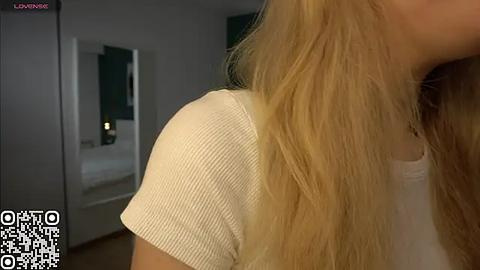 Media: Video of a woman with long blonde hair, wearing a white ribbed tank top, captured from the side. Background shows a dimly lit room with a green wall and a mirror. QR code in the bottom left corner.