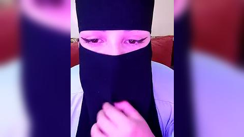 Media: A video of a veiled woman with long eyelashes, wearing a black headscarf, seated on a red couch with a blurred background.