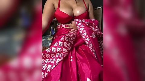 Media: Video of a woman with large breasts, wearing a red bra and a red and white patterned sarong. She is seated, her body slightly angled, with a blurred background.