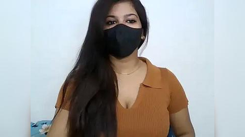 Media: Video of a South Asian woman with long black hair, wearing a black mask, mustard-colored ribbed top, and a gold necklace, indoors against a white wall.