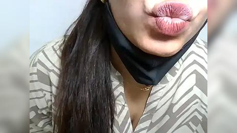 Media: Video of a woman with long dark hair, wearing a black mask, puckered lips, and a light-colored patterned blouse. Background is blurred, focusing on her face.