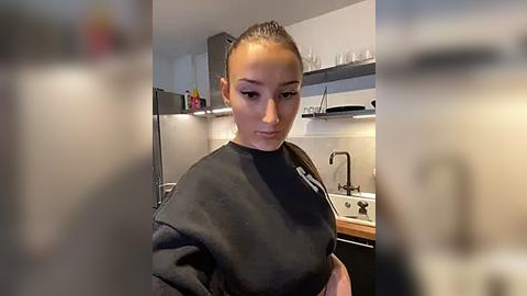 Media: Video of a young woman with light skin and straight black hair in a ponytail, wearing a black t-shirt, standing in a modern kitchen with stainless steel appliances, white countertops, and glass shelves.